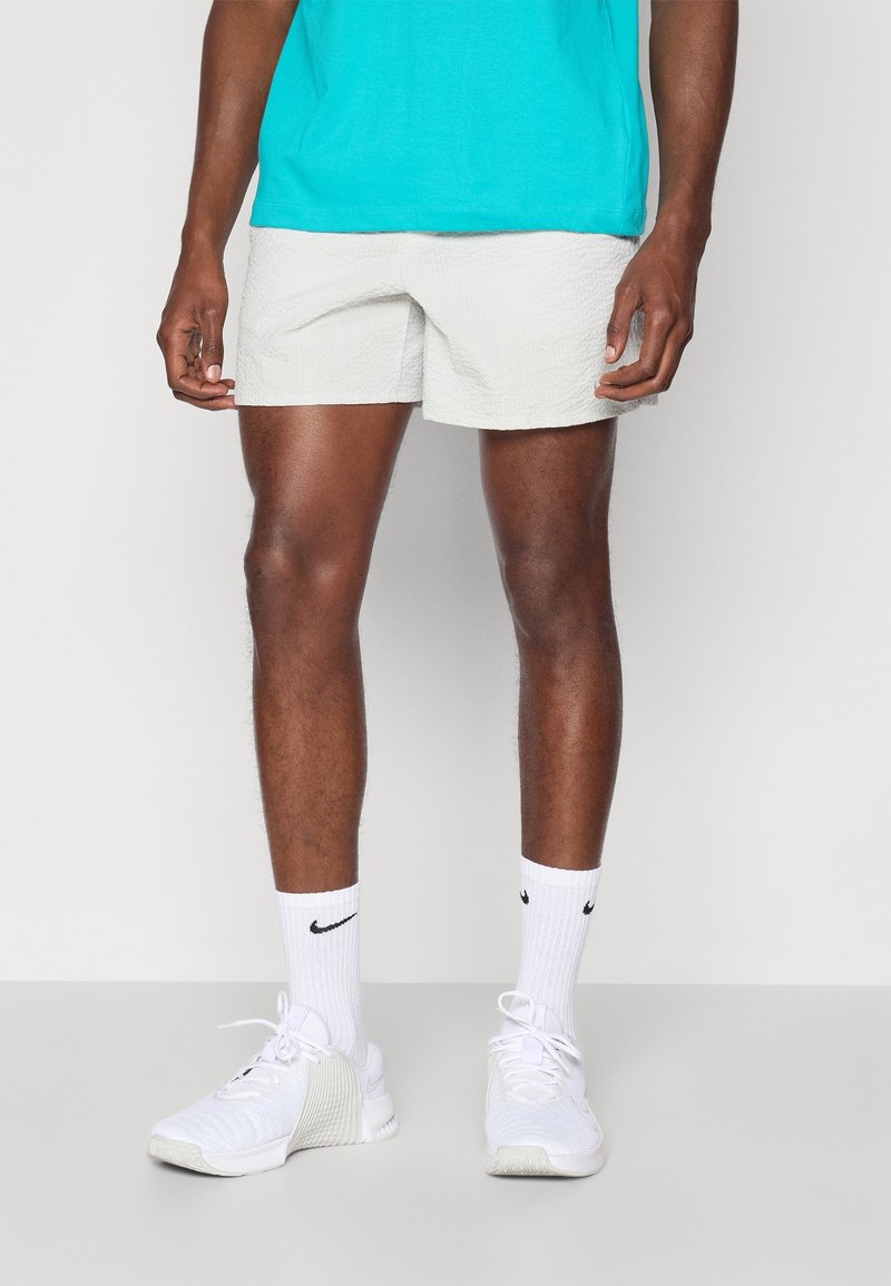 Homme portant un short gris clair texturé, une chemise turquoise, des chaussettes blanches Nike et des chaussures de sport blanches, debout devant un fond uni.
