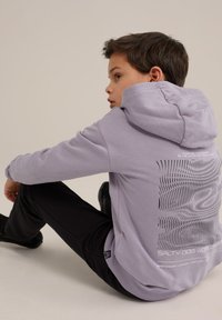 Fialová mikina s textúrovaným grafickým dizajnom na zadnej strane, kombinovaná s čiernymi nohavicami. Materiál vyzerá mäkko a neformálne, s voľným strihom.