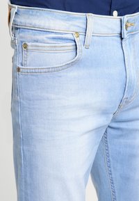 Jeans de mezclilla azul claro con un diseño clásico de cinco bolsillos, herrajes en tono dorado y un desgaste sutil en el muslo y las caderas.