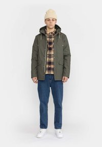 Olivgrüne Jacke mit Kapuze, Fronttaschen und Reißverschluss. Über einem karierte Hemd getragen, kombiniert mit blauen Jeans und weißen Sneakers.