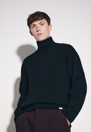 Ung mand med krøllet hår iført en sort højhalset sweater og mørke bukser, stående mod en ensfarvet lys baggrund.