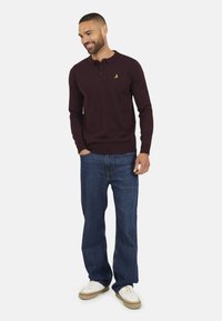 Polo a maniche lunghe color bordeaux con colletto a tre bottoni e piccolo logo giallo. Abbinato a jeans blu e sneakers bianche.