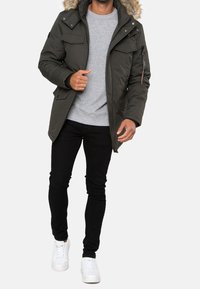 Oliwkowo zielona kurtka parka z kapturem podszytym futrem, wieloma kieszeniami z przodu oraz kieszenią zapinaną na suwak na rękawie, noszona na szarej bluzie i czarnych dżinsach.