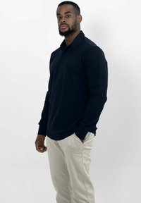 Marineblauwe polo met lange mouwen en een kraag, voorzien van een drieknopen sluiting en geribbelde manchetten, gecombineerd met lichtbeige broeken.