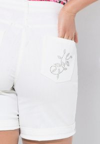 Shorts de mezclilla blanca con puños enrollados, que presentan un bolsillo trasero decorado con un diseño floral de pequeños strass.