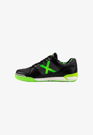 Zapatilla deportiva negra con suela verde, "X" verde en el lateral, cordones negros y forro interior de camuflaje verde, vista desde el lado exterior.