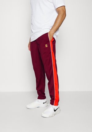Homme portant un pantalon de survêtement bordeaux avec une bande rouge et un liseré blanc, des baskets blanches Nike et un t-shirt blanc, debout les mains dans les poches.