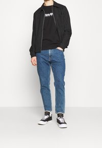Svartrandig zip-up jacka över en svart grafik T-shirt, ihop med blå jeans och svartvita sneakers.