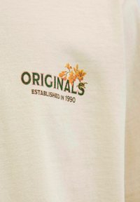 Krém színű, pamutból készült póló, zöld "ORIGINALS" felirattal és sárga virágos hímzéssel, mellette az "ESTABLISHED IN 1990" felírat.