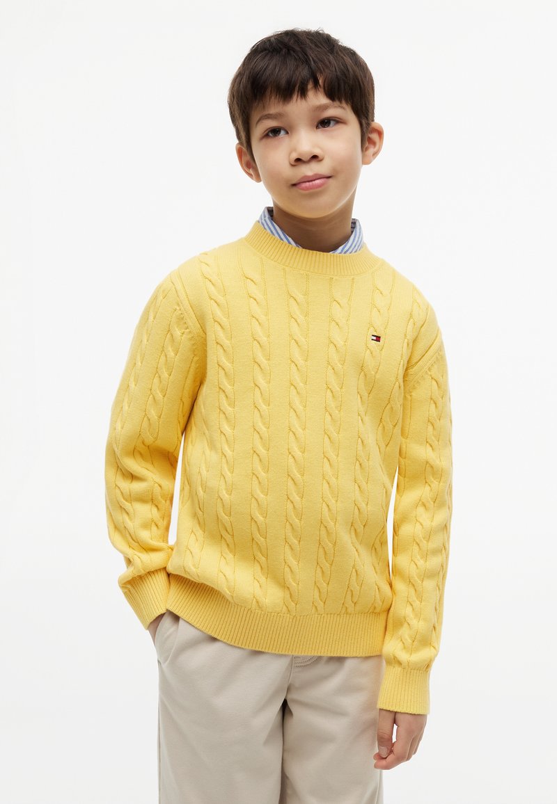 Garçon portant un pull jaune torsadé sur une chemise rayée bleue et un pantalon beige, debout les mains dans les poches sur fond blanc.