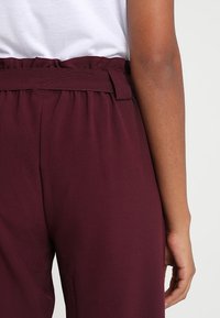 Bordeaux kleurige broek met een geraffineerde taille, voorzien van een brede tailleband en lussen voor een riem, met een gladde textuur en een losse pasvorm.