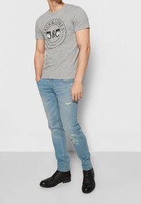 Camiseta gris de manga corta con un logo circular negro, combinada con jeans desgastados de color azul claro y botas negras con cordones.