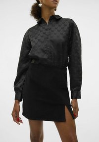 Camisa negra con botones y un sutil patrón de lazos, de mangas largas y con cuello, combinada con una mini falda de pana negra con una abertura lateral.