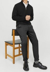 Maglione nero con zip, pantaloni grigi a quadri, scarpe nere con superficie texturizzata, seduto su una sedia di legno con seduta nera.