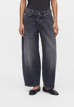 Person iført vidde-sort falmede jeans med matchende stofbælte bundet i taljen og sorte flade sko.