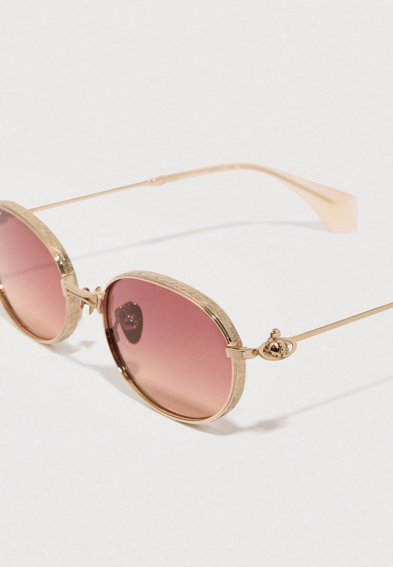 Ovale solbriller med pink gradient linser, guldtonet metal stel, strukturerede detaljer på stellet og dekorativt beslag på hængslerne.