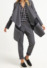 Mujer vestida con un abrigo gris de apertura frontal, blusa a cuadros en blanco y negro, pantalones grises con cordón y zapatos negros de tipo slip-on.