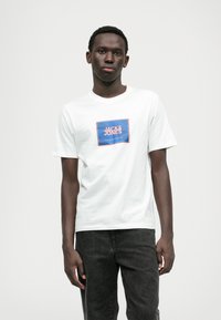 Biela bavlnená tričko s modrým obdĺžnikovým logom, ktoré obsahuje nápis "JACK & JONES" v bielej a červenej farbe. Krátke rukávy, voľný strih.