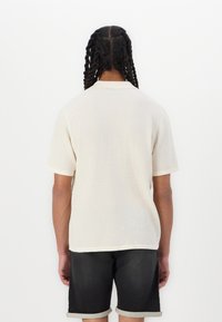 Short-sleeve beige polo shirt with a textured knit fabric, featuring a contrasting collar and hem. Worn with rolled-cuff dark shorts.