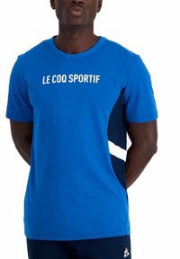 Camiseta de manga corta azul con texto en blanco "LE COQ SPORTIF". Presenta paneles de acento en color marino y un diseño de cuello redondo. Textura de material suave.