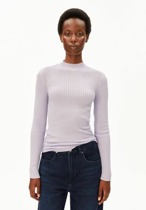 Frau mit kurzen lockigen Haaren, die ein hellviolettes, geripptes Langarmoberteil und dunkelblaue Jeans trägt und vor einem schlichten weißen Hintergrund steht.