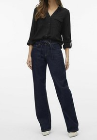 Blusa nera con bottoni abbinata a jeans in denim scuro a vita alta e gamba larga. Le caratteristiche principali includono maniche arrotolate e cuciture a contrasto sui jeans.