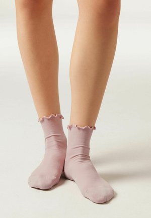 Nohy v světle růžových žebrovaných kotníkových ponožkách s volánkovými lemy stojící na jednobarevném bílém povrchu.