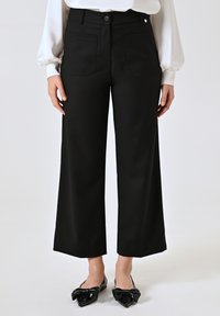 Pantaloni neri a gamba larga con una texture liscia, dotati di tasche frontali e posteriori, e chiusura con bottone. Indossati con ballerine nere a punta.