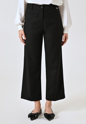 Pantaloni neri a gamba larga con una texture liscia, dotati di tasche frontali e posteriori, e chiusura con bottone. Indossati con ballerine nere a punta.