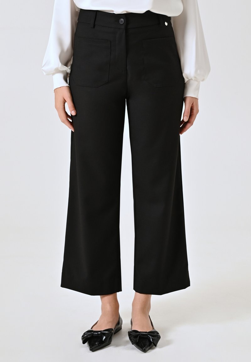 Pantaloni neri a gamba larga con una texture liscia, dotati di tasche frontali e posteriori, e chiusura con bottone. Indossati con ballerine nere a punta.