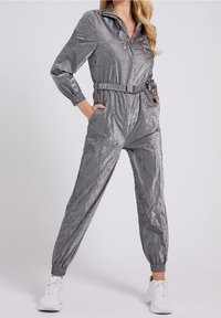 Mono metálico gris con cremallera frontal, cintura elástica y piernas con puños. Cuenta con dos bolsillos frontales y una cintura con cinturón para ajustar.