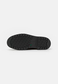 Suela de zapato de goma negra con un diseño texturizado que presenta profundos surcos y un acento verde. Incluye el logo de la marca y un diseño de patrón destacado.