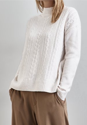 Femme portant un pull blanc à col roulé en maille torsadée avec un ourlet côtelé, associé à un pantalon marron, mains dans les poches, fond neutre.