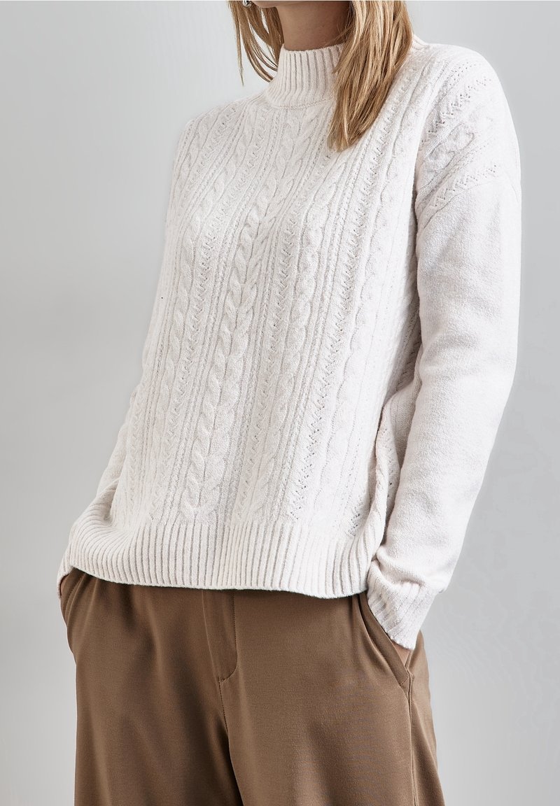 Femme portant un pull blanc à col roulé en maille torsadée avec un ourlet côtelé, associé à un pantalon marron, mains dans les poches, fond neutre.