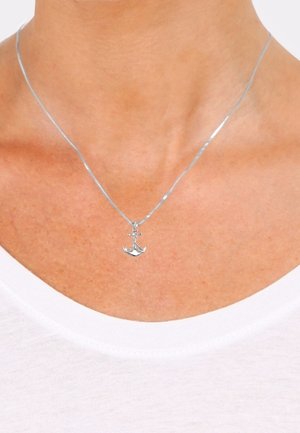 Pendentif ancre argentée sur une chaîne fine portée autour du cou au-dessus du col d'une chemise blanche.