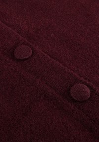 Bordeaux wollen stof met een gestructureerd oppervlak, voorzien van twee ronde gebreide knopen langs een naad. Het geheel straalt zachtheid en warmte uit.