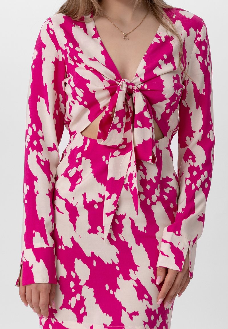 Femme portant une robe à manches longues aux motifs roses vifs et blancs avec un nœud à l'avant, dévoilant une petite découpe au niveau du ventre.