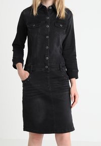 Svart denimklänning med lång ärm, två bröstfickor, framknäppning med knappar och dekorativa nitar på fickorna. Slät textur.