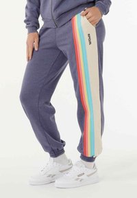 Persoon in paarse joggers met regenboogstrepen en de tekst "RIP CURL" op de zijkant, gecombineerd met witte sneakers en witte sokken.