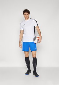 Nike sportset met een witte shirt met zwarte accenten, blauwe shorts en zwarte sokken. De speler houdt een gestreepte voetbals in zijn hand.
