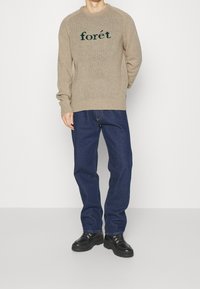 Beżowy sweter z dzianiny z napisem "forêt" w ciemnozielonym kolorze, zestawiony z niebieskimi dżinsami i czarnymi sznurowanymi butami. Sweter ma ściągacze na mankietach i dolnym brzegu.