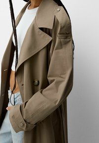Beige trenchcoat med bred krage, dubbla knappar, knapprad och en avslappnad silhuett, parat med en vit topp och ljus denim.