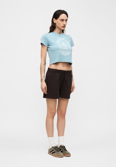 Νεαρή γυναίκα με μακριά σκοτεινά μαλλιά στέκεται μπροστά, φοράει γαλάζιο κοντό τοπ, μαύρα σορτς, λευκές κάλτσες και καφέ αθλητικά παπούτσια.