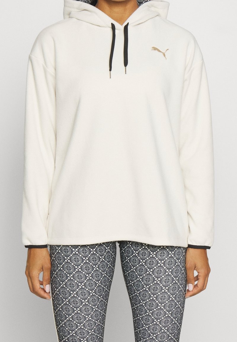 Personne portant un sweat à capuche blanc Puma avec des cordons noirs et des leggings noirs et blancs à motifs visibles des épaules aux hanches.