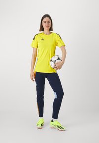Geltona sportinė marškinė su juodais akcentais, tamsiai mėlynos kelnės su daugiakuliniais šoninėmis juostomis ir ryškiai geltonos futbolo bateliai. Laiko futbolo kamuolį.