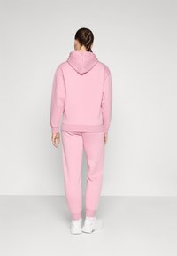 Różowy dres składający się z bluzy z kapturem i dopasowanych spodni dresowych. Materiał wydaje się miękki i ma luźny krój. Widać białe sneakersy.