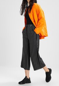 Oranje gewatteerde jas, zwarte top, zwart-wit gestreepte wijde broek en zwarte sneakers met vetersluiting. Over het geheel een gladde textuur.