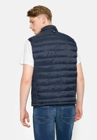 Marinefarbener Steppweste mit hohem Kragen, horizontaler Steppung und Druckknopfverschluss am Hals, kombiniert mit einem weißen T-Shirt und Jeans.