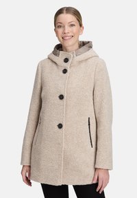 Beige fleece jas met capuchon, zwarte knopen, zijzakken met zwarte accenten en een zachte textuur. Recht gesneden model, halflang.