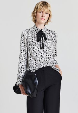 Schwarz-weiß gemusterte Bluse mit Halsband, die ein geometrisches Design aufweist. Kombiniert mit schwarzen, hoch taillierten Hosen und einer glänzenden schwarzen Clutch.